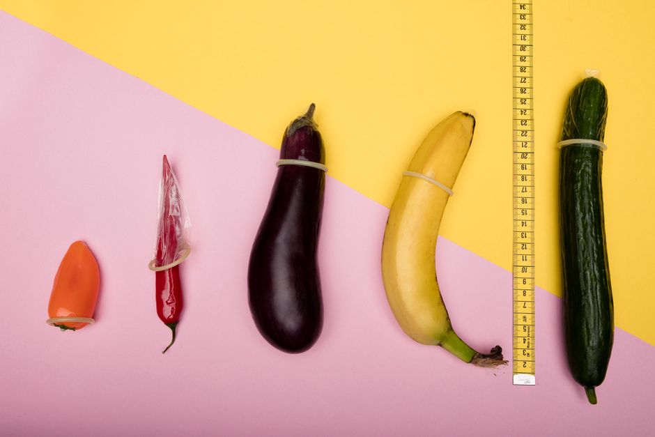 A staged arrangement of five vegetables and fruits laid out on a split-colored background with yellow and pink sections, measuring from left to right. The first item is a small orange bell pepper, smooth and glossy with a slight curve. Next, a red chili pepper wrapped in a clear plastic band, with a shiny surface and a pointed tip. The third item is a large, dark purple eggplant with a smooth, glossy skin, slightly curved with a green stem at the top. Following is a ripe, yellow banana with a slight bend and a smooth textured peel, also secured with a plastic band. The last item is a long, dark green cucumber with a bumpy surface, attached to a measuring tape placed vertically beside it, indicating its length. The background provides a clean, minimal setting emphasizing the fresh produce, suitable for content related to gardening or outdoor produce cultivation, with natural lighting highlighting the textures and colors of the vegetables and fruit.