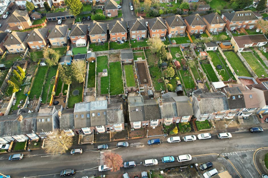 An aerial view of a row of terraced houses in Ruislip with well-maintained gardens, showing various landscaping and garden maintenance styles. The gardens feature lush green lawns, flower beds, paved pathways, and a range of plants and shrubs, some with trimmed hedges while others have more natural, leafy borders. Several gardens display outdoor furniture, toy trampolines, and storage sheds, indicating residential outdoor use. The roofs of the houses are shown with roofing tiles, and the street in front has parked cars. Trees are visible along the sidewalks and within some gardens, providing shade and greenery to the scene. The overall setting reflects typical suburban garden care and landscaping efforts, supported by services such as hedge trimming and lawn care provided by Gardeners Ruislip.