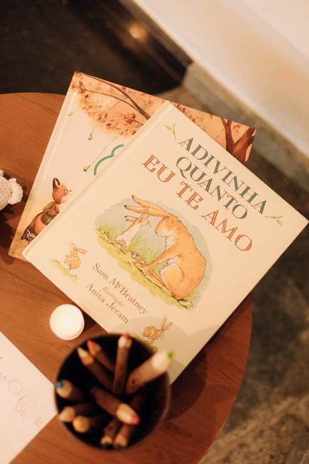 A close-up view of a wooden table outdoors, featuring two children's books with colorful illustrations of a fox and forest animals, titled 'Adivina Quánto Te Amo.' The books are slightly angled, revealing their engaging covers. In front of the books, there is a small white candle or decorative orb, and in the foreground, a round container filled with various colored pencils and crayons, some sharpened. The setting appears to be a garden or patio area with natural lighting, creating a cozy and inviting atmosphere suitable for outdoor reading or artistic activities.